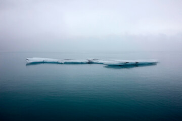 Naklejka premium Isolated minimalist ice floe floating in calm turquoise glacial lagoon with thick fog in horizontal Iceland view