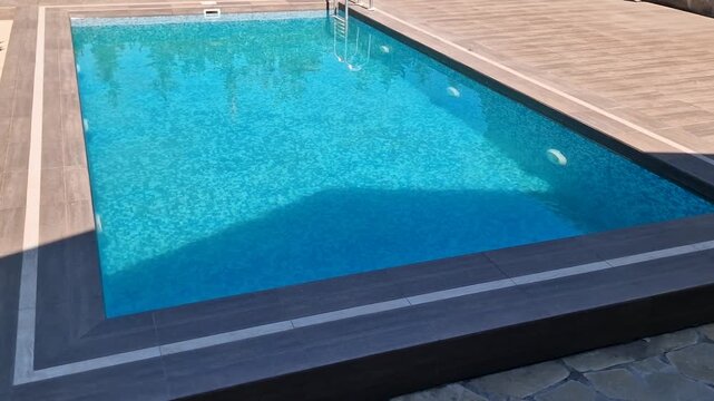 Rectangular outdoor swimming pool with crystal clear blue water, mosaic tile bottom, and a modern wooden texture tiled deck on a sunny day. Luxury backyard or resort area