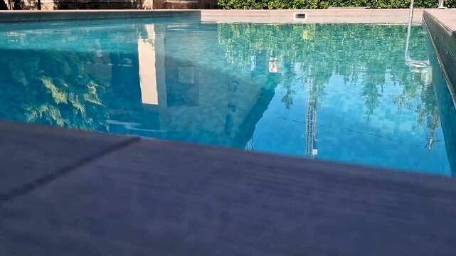 Rectangular outdoor swimming pool with crystal clear blue water, mosaic tile bottom, and a modern wooden texture tiled deck on a sunny day. Luxury backyard or resort area