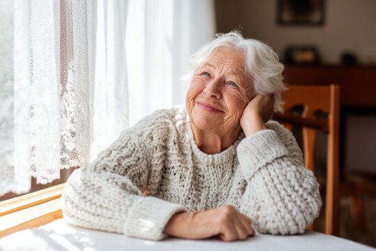 The elderly woman daydreaming by a sunlit window in a cozy knitted sweater