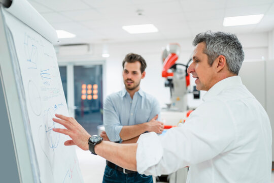 Mature engineer explaining business plans to young man at white board in factory