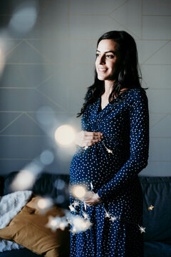 Thoughtful pregnant woman looking away while standing with hands on stomach in living room