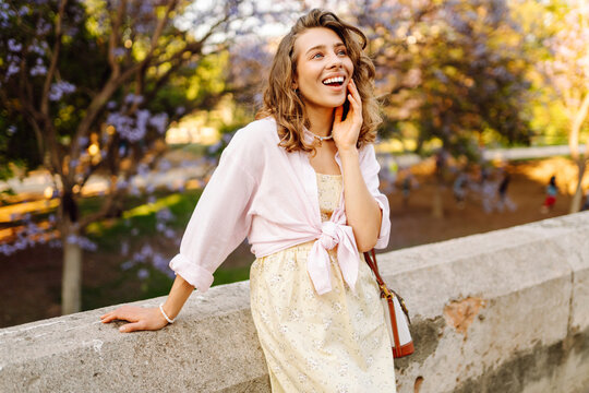 A young woman strolls along a path in a park at sunset. A beautiful woman in a dress enjoys a sunny day near blossoming trees. Concepts of beauty, style, and pleasure.