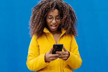 Surprised woman with mouth open using mobile phone while standing against blue wall