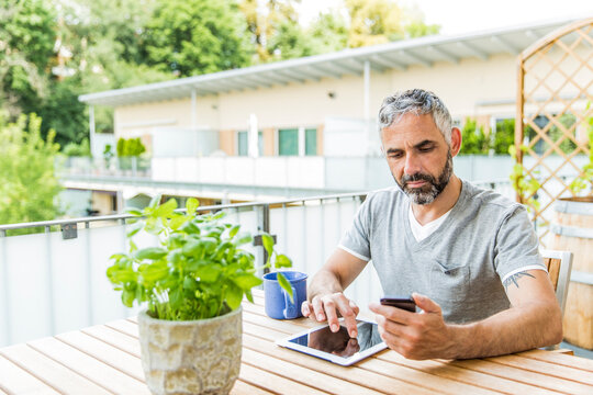 Portrait of man sitting on his balcony using smartphone and digital tablet
