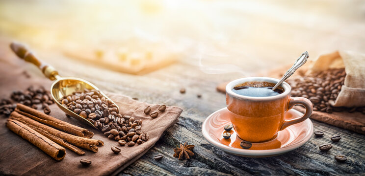Cup of hot black coffee on a rustic wooden table surrounded by roasted coffee beans, cinnamon sticks, and star anise. 