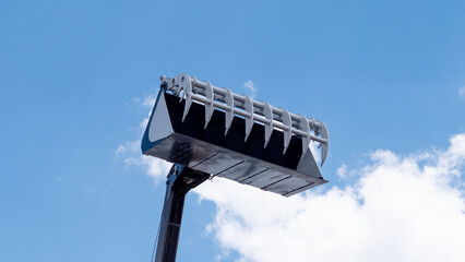 A large excavator bucket is lifted high against a clear blue sky with soft white clouds, showing the metal teeth and industrial equipment from a low angle © bisonov