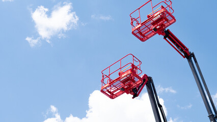 Two red elevated work platforms on extended booms are shown from a low angle against a bright blue sky with white clouds, creating an industrial maintenance scene with open space © bisonov