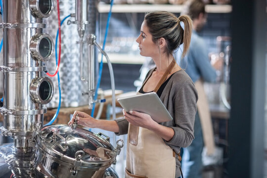 Young woman with digital tablet looking at machine in distillery