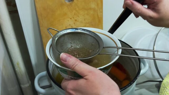 Chef hand straining ladled broth through sieve, close-up