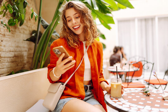 Smiling woman with phone sits at table in cafe with soft drink. Woman enjoys coffee on the cafe terrace with a phone in her hand. Concept of relaxation, drinks, and weekends.