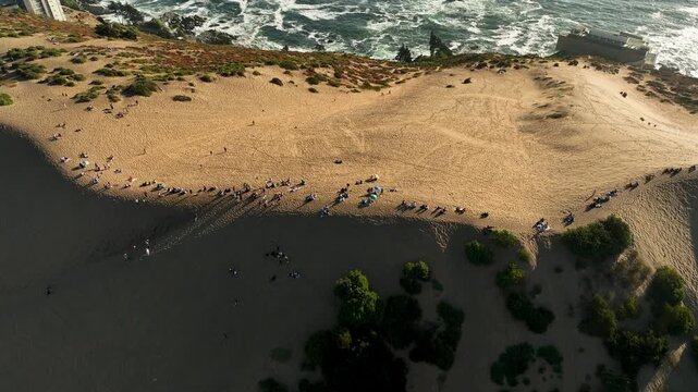 Drone forward flight with gimbal tilt down transition from high angle to vertical top-down view over people gathered on sand dune ridge at sunset in Concon Chile