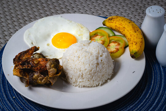 close up photo of danggit breakfast or sun dried rabbit fish with sunny side up egg, fried rice, tomatoes and cucumber with a piece of ripe banana on a white circular plate.