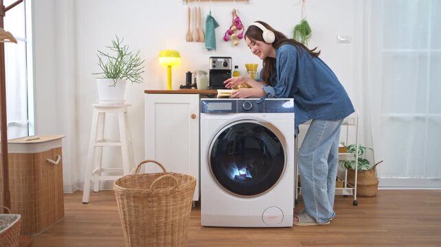 Young Asian woman checking the progress of a washing machine cycle while listening to music on the top of laundry machine