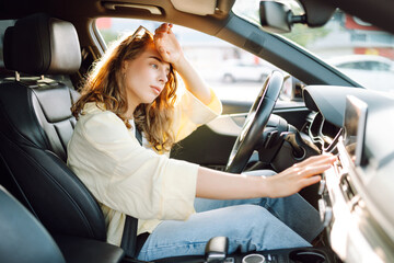 A young woman sits behind the wheel of a hot car. A female driver feels unwell due to the heat in the car on a sunny day. Concept of air conditioning, cooling, and transportation.