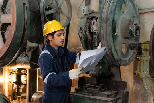 Asian industrial technician in hardhat checking paperwork next to heavy factory machinery.