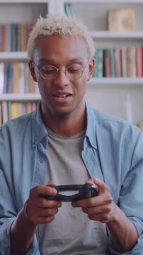 A joyful young African American man with blonde hair and glasses plays video games. He holds a gamepad, laughs, and expresses excitement while gaming in a cozy library home setting.