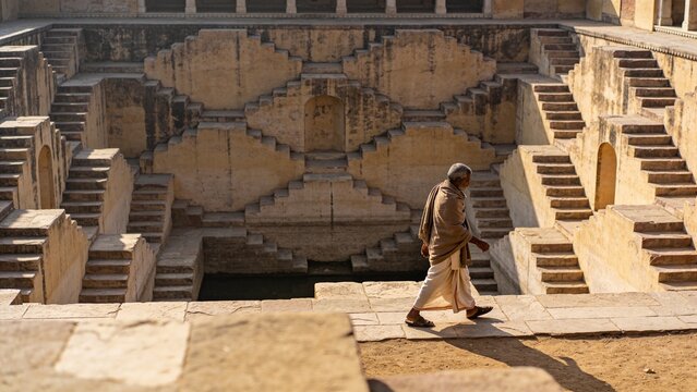 Walking senior man wearing traditional shawl along sunlit stepwell ledge, stone steps, copy space