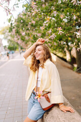 Naklejka premium A smiling woman enjoys a sunny day in a park, surrounded by blooming trees. The woman, in casual clothes, poses outdoors. Concept of beauty, fashion, and relaxation.