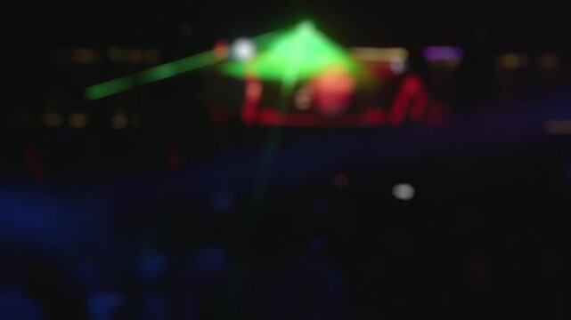 Out-of-focus background of a crowded nightclub with green laser beams and colorful stage lights illuminating the dj and the dancing audience, creating an energetic and vibrant atmosphere