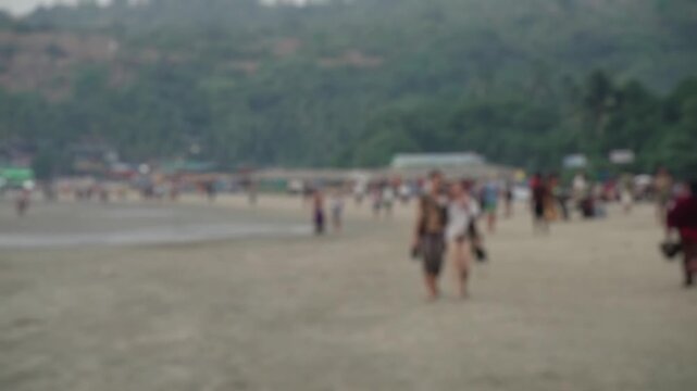 Out of focus shot of a crowd of unrecognizable people walking on a sandy beach during the day, with a lush green hill in the background, creating a dreamy and anonymous atmosphere