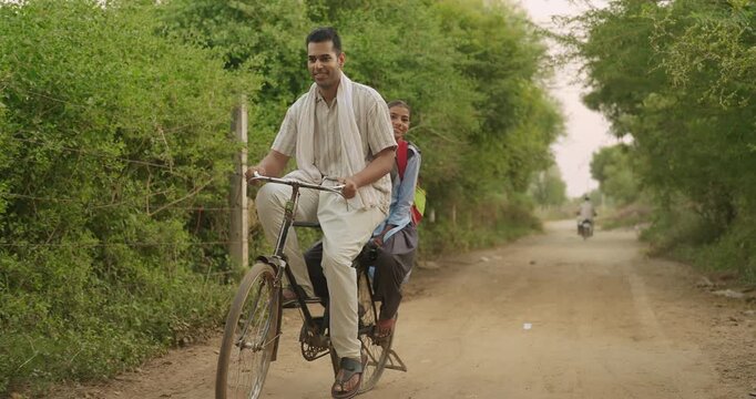 Indian happy young adult dad talk speak drop school to teen girl child rural area place road path way. Smiling female wear uniform with bag sitting on cycle back seat enjoy ride have fun joy desi life