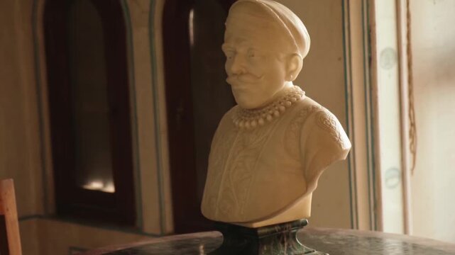 Elegant marble bust of a historical Indian figure, possibly a maharaja, displayed inside a traditional palace, with soft light coming from a window illuminating the intricate details