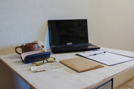 A side perspective of a white desk with a laptop, a clipboard with paper and a pen, a craft notebook, glasses with a case, a microfiber cloth, a brown cup, and a skull figurine
