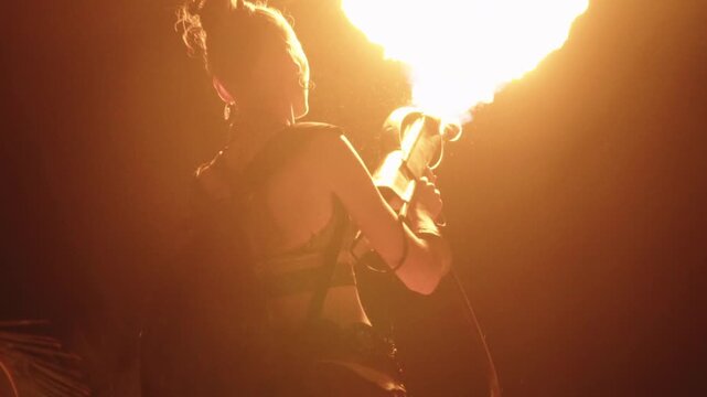 Female artist performing a dangerous fire show with a flamethrower at night, creating a powerful burst of orange flames and smoke against the dark background during a festival