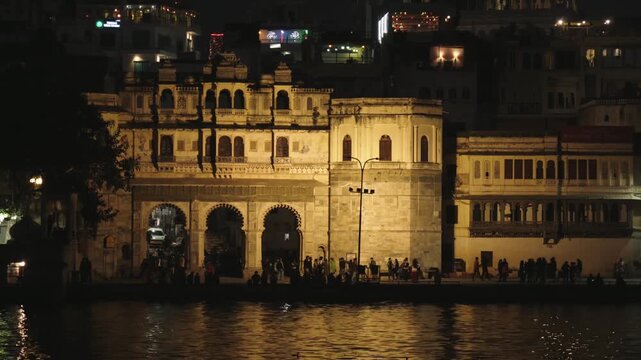 Beautifully illuminated architecture of the Gangaur Ghat in Udaipur, India, reflecting on the tranquil waters of Lake Pichola at night, with silhouettes of people enjoying the evening