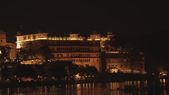 Beautiful view of the illuminated City Palace complex at night, with its majestic facade and warm lights reflecting on the tranquil waters of Lake Pichola in Udaipur, Rajasthan, India