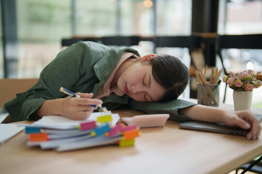 Young Asian woman sleeping at her office desk, surrounded by documents and a pen, illustrating modern workplace stress, exhaustion, burnout, and an overwhelming workload
