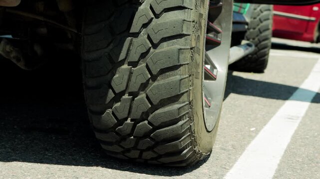 Detailed shot of heavy duty all terrain tire shows deep grooves and block pattern. Large rubber wheel mounted on vehicle rim with subtle red accent. Paved surface of parking space has visible white li