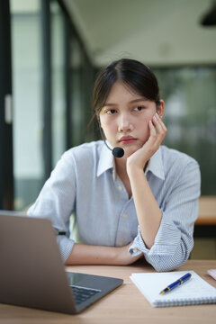 Asian woman feeling burnout during work, resting her chin on her hand, looking bored and tired while wearing a headset and sitting at her desk with a laptop and notebook