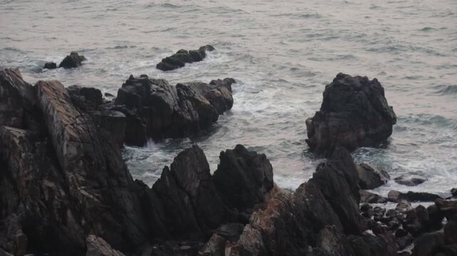 Overcast seascape with dramatic ocean waves breaking against the sharp, dark rocks of a rugged shoreline, creating white foam and spray in a powerful and relentless natural display
