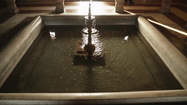 Small ornate indoor fountain flowing in a rectangular marble basin filled with wish coins, creating ripples on the water surface and illuminated by a bright ray of sunlight from a window