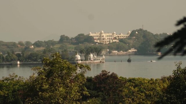 Panoramic view of the historic Jag Mandir island palace and the City Palace on the banks of Lake Pichola, with tourist boats sailing on the tranquil water on a hazy summer day in Udaipur, India