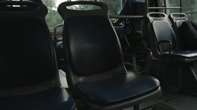 Empty blue seats inside a public transport bus moving along a tree-lined road, with sunlight casting shadows across the interior and the landscape passing by the window