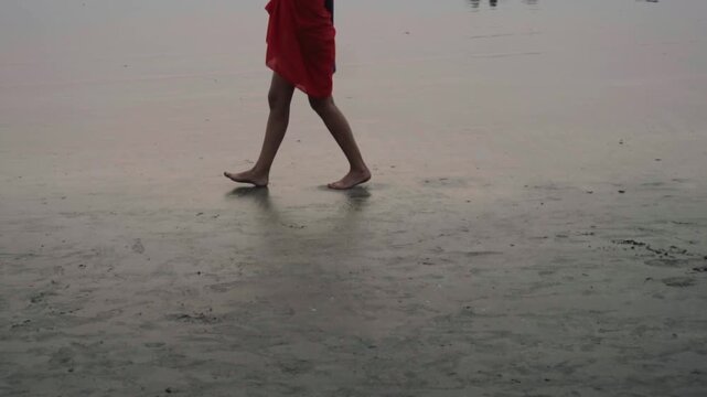 Lower body shot of a woman wearing a red skirt walking barefoot on the wet sand of a beach, her reflection visible on the damp surface as she strolls along the shoreline