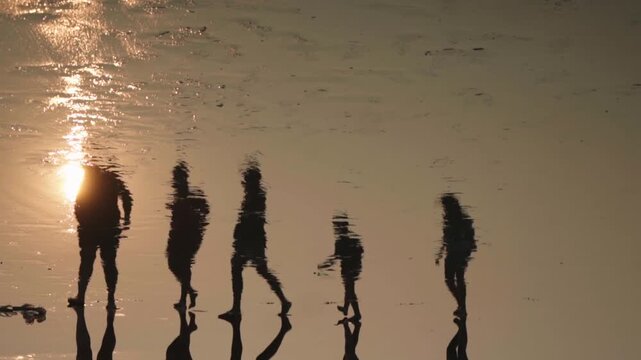 Silhouettes of a group of people walking on the wet sand of a beach at sunset, with their dark figures and the golden sunlight beautifully reflected on the shimmering water surface