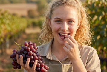 Fototapeta premium Young woman happily eating fresh red grapes, holding a bunch of grapes in a vineyard