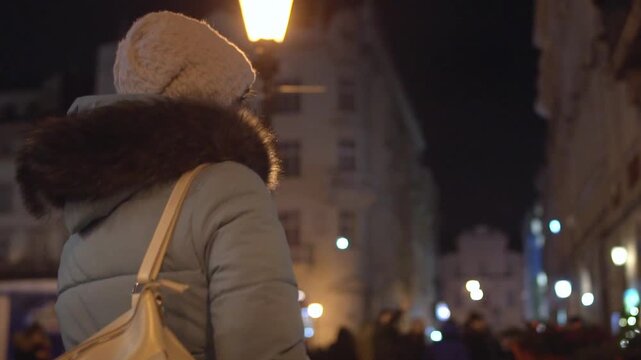 Person walks from behind, wearing warm winter clothing, including knitted hat and fur-trimmed coat, carrying shoulder bag. Street lights and holiday decorations glow, creating festive atmosphere in ur