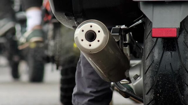 Detailed shot of shiny metal exhaust pipe and treaded rubber rear wheel of motorcycle. Slight smoke or vapor rises from exhaust, suggesting active engine. Background shows numerous other motorcycles a