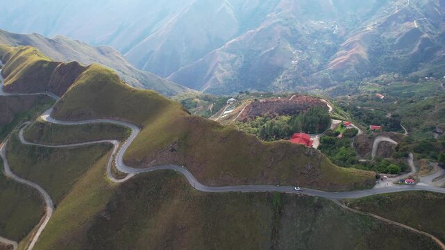 Cinematic aerial view winding mountain road cutting through rolling green highlands and rugged valleys. Scenic switchbacks, remote countryside buildings, and dramatic mountain terrain