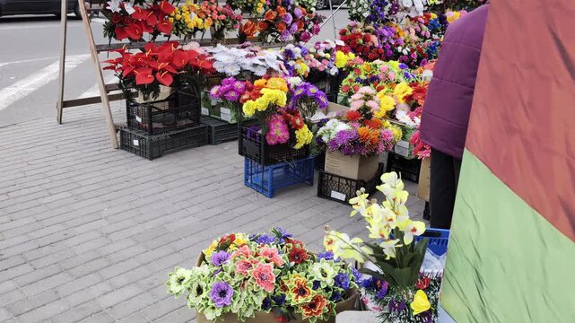 Vibrant assortment of fake flowers arranged in crates and boxes on grey brick sidewalk. Many different hues of red, yellow, purple, and white blooms visible. Partial view of person at market stall. Ra
