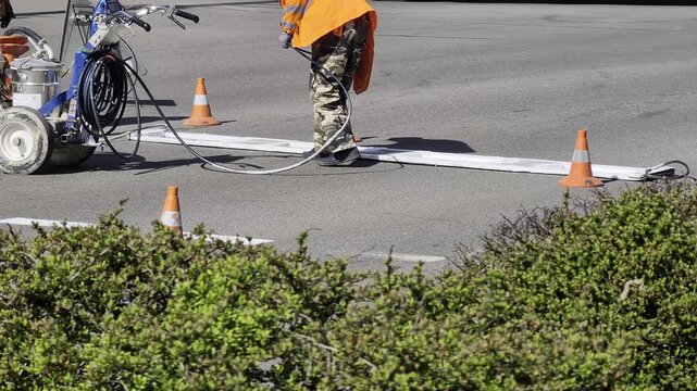 Outdoor scene showing road worker applying white traffic line paint on asphalt street, utilizing line striper machine and orange safety cones. Public service maintenance activity for road infrastructu