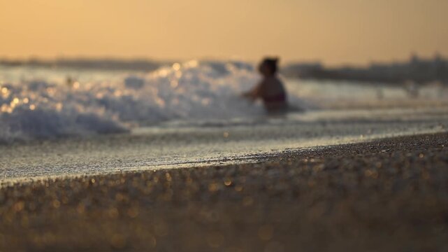 View of one person enjoying calming movement of ocean water, wading in gentle waves with golden hour light reflecting on surface. Sand and water close up with bright bokeh, background is blurry. Vacat