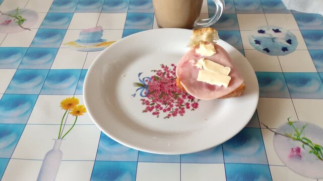Hand taking ham sandwich from plate on patterned tablecloth with coffee mug in background, first person view