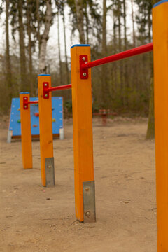 Close-up view of a row of bright yellow wooden posts connected by red metal parallel bars in a sandy playground clearing. Background shows scattered trees