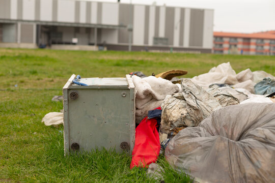 A pile of discarded household items and rubbish scattered on a grassy lot in an urban area. The foreground shows a small white appliance tipped on its side